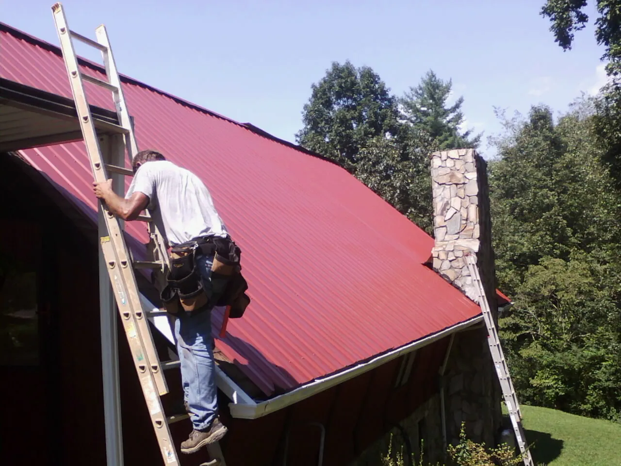 Roofer on a ladder beside a red metal roof and stone chimney — Soffit & Fascia Repair by Roofing Brothers in Crabtree